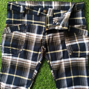 Op de afbeelding: Een blauwe, bruine en witte geruite broek met een ritsvlieg en een knoopsluiting. De broek heeft twee grote zakken aan de voorkant.