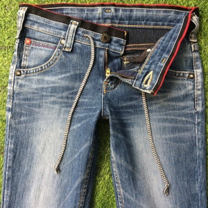 May include: Blue denim jeans with a drawstring waist and a zip fly. The jeans have a classic five-pocket design with red stitching details and a black waistband with red trim. The jeans are laid flat on a green surface.