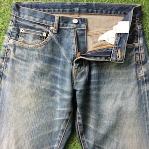 Puede incluir: Un par de pantalones vaqueros azules de mezclilla con cierre de cremallera y botón. Los vaqueros tienen un aspecto descolorido y desgastado, con signos visibles de uso.