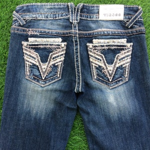 Può includere: Un paio di jeans blu denim con tasche posteriori decorate. Le tasche presentano un design ricamato bianco e argento a forma di V. I jeans hanno un'etichetta "VIGOSS" sul retro della cintura.