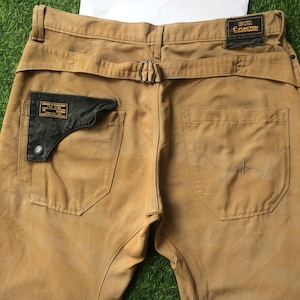 May include: A pair of brown cargo trousers with a black leather patch on the left pocket and a black label with the text "Edwin & Function" on the right pocket. The trousers have a belt loop and a buckle closure.