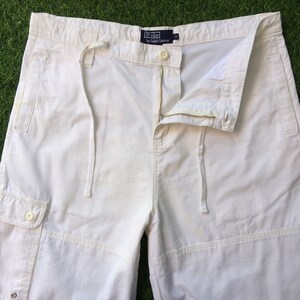 May include: White cargo pants with a drawstring waist and a zipper fly. The pants have a patch pocket on the left leg and a label that reads "Polo by Ralph Lauren".