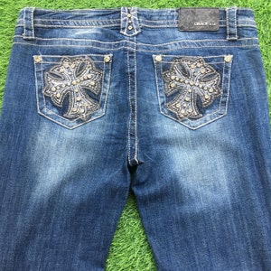 Puede incluir: Un par de pantalones vaqueros azules de mezclilla con un diseño de cruz en los bolsillos traseros. Los vaqueros tienen un lavado oscuro y un aspecto desgastado. La etiqueta en la parte posterior de la cintura dice "GRACE in LA".