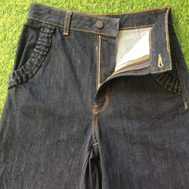 May include: Dark blue denim jeans with a zipper fly and a ruffled detail on the front pockets.