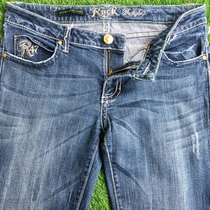 Op de afbeelding: Een paar blauwe denim jeans met de tekst "Rock by Wrangler" op de tailleband en "R47" geborduurd op de voorzak.