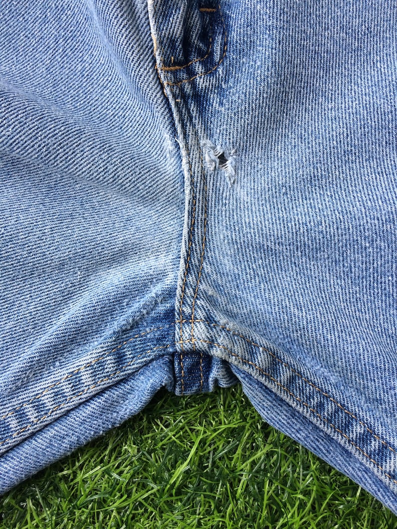 Pode incluir: Close-up de um par de cal&ccedil;as de ganga azuis com um pequeno furo perto da costura. As cal&ccedil;as est&atilde;o sobre um leito de relva verde.