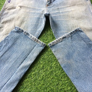 May include: A pair of light blue denim jeans with faded areas and a distressed look. The jeans are laid out on a green grassy surface.