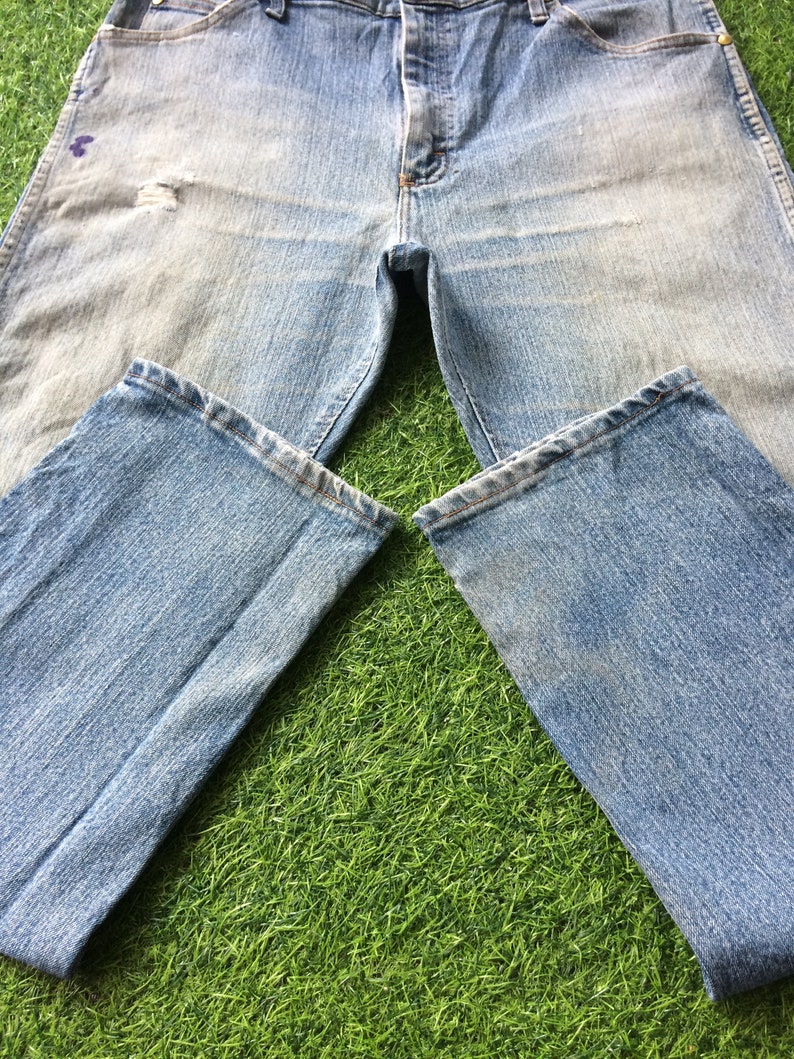 May include: A pair of light blue denim jeans with faded areas and a distressed look. The jeans are laid out on a green grassy surface.