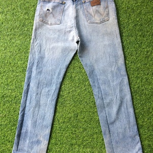 May include: A pair of light blue denim jeans with a faded look. The jeans have a brown leather patch on the back with the word "Wrangler" printed on it.