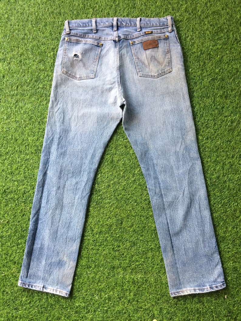 May include: A pair of light blue denim jeans with a faded look. The jeans have a brown leather patch on the back with the word "Wrangler" printed on it.
