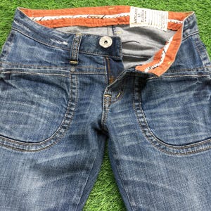 Puede incluir: Primer plano de unos vaqueros de mezclilla azules con un diseño de bolsillo único. Los vaqueros tienen cierre de botón y cremallera. La cintura presenta un forro a rayas naranja y blanco. Los vaqueros están sobre una superficie verde.