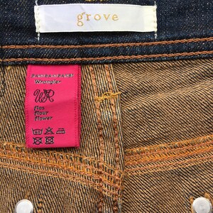 May include: Close-up of denim jeans with a brown and blue wash. The jeans have a white tag with the word "grove" in gold lettering. A pink tag reads "RAN/BAN/RANBI Wrangler" and features the letters "WR".