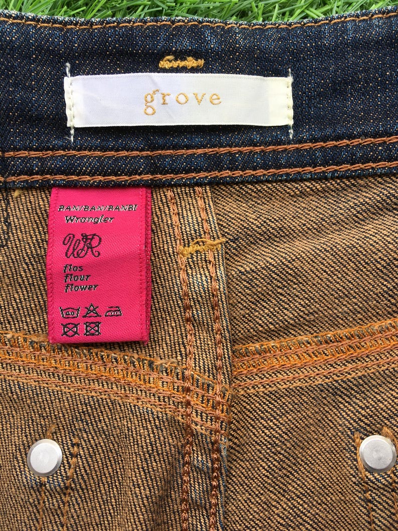May include: Close-up of denim jeans with a brown and blue wash. The jeans have a white tag with the word "grove" in gold lettering. A pink tag reads "RAN/BAN/RANBI Wrangler" and features the letters "WR".
