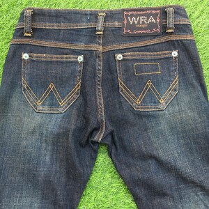 May include: Dark wash denim jeans with a classic five-pocket design. Features include a W-shaped stitch on the back pockets, copper-colored stitching, and a WRA Wrangler label. The jeans are displayed on a green surface.