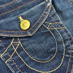 May include: Close-up of a section of blue denim jeans. The jeans feature a pocket with decorative stitching in a contrasting yellow thread. A gold button with the text "JEANS TRUE RELIGION BRAND" is visible.