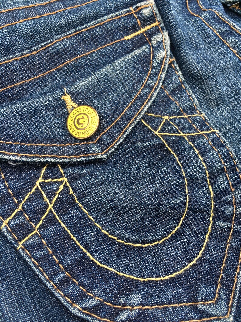 May include: Close-up of a section of blue denim jeans. The jeans feature a pocket with decorative stitching in a contrasting yellow thread. A gold button with the text "JEANS TRUE RELIGION BRAND" is visible.