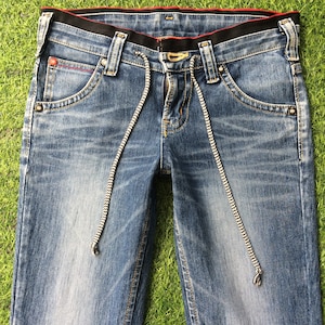 May include: Close-up of blue denim jeans with a black waistband and red stitching details. The jeans feature a drawstring with a black and white pattern. The pockets have a unique design, and the jeans are laid on a green surface.