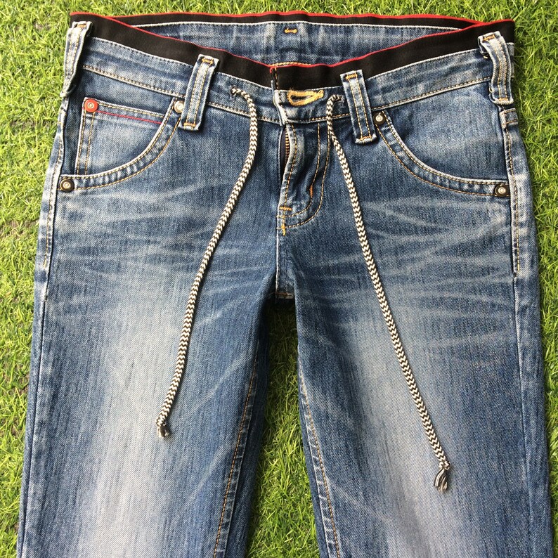 May include: Close-up of blue denim jeans with a black waistband and red stitching details. The jeans feature a drawstring with a black and white pattern. The pockets have a unique design, and the jeans are laid on a green surface.