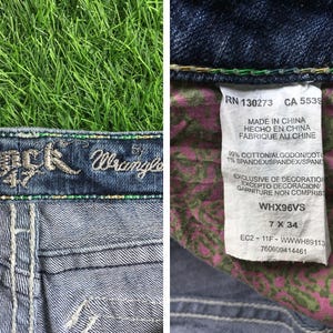 Op de afbeelding: Blauwe denim jeans met een Rock & Wrangler logo op de tailleband. De jeans heeft een bloemenprint op de achterzakken en een label met de tekst "RN 130273 CA 5539 Made in China Hecho en China Fabrique au Chine 99% Cotton/Algodon/Coto 1% Spandex/Spandex/Spand Exclusive of Decoration Excepto Decoracion/ Garniture Non Compris WHX96VS 7 X 34 EC2-11F-WWWH89113 760609414461".