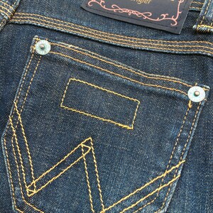 May include: Close-up of dark blue denim jeans with a back pocket featuring the Wrangler logo stitched in yellow thread. The jeans have a dark blue label with the Wrangler logo and decorative stitching.