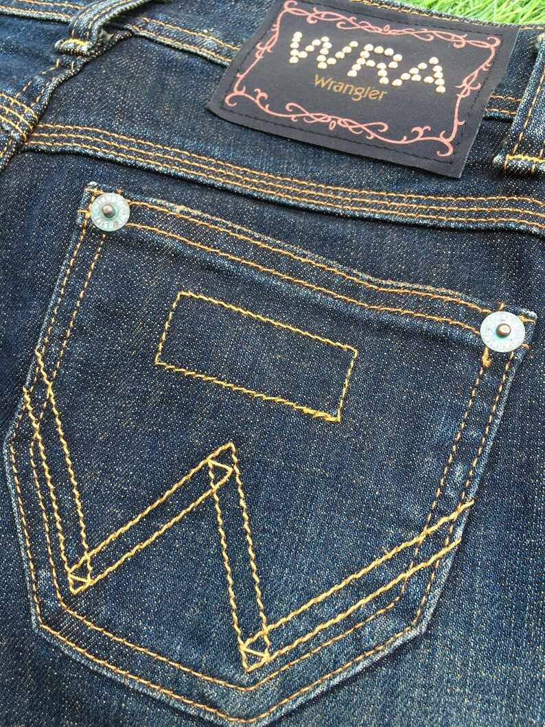May include: Close-up of dark blue denim jeans with a back pocket featuring the Wrangler logo stitched in yellow thread. The jeans have a dark blue label with the Wrangler logo and decorative stitching.
