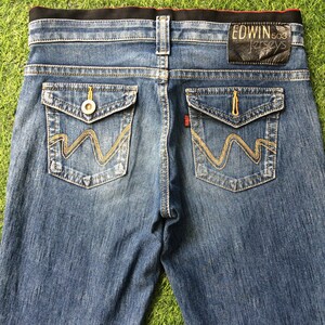 May include: Blue denim jeans with a black waistband and two back pockets. The pockets feature a distinctive design with gold stitching. The jeans display the brand name "EDWIN" and "503 Jerseys" on a black leather patch.