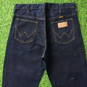Peut inclure: Un jean en denim bleu fonc&eacute; avec un &eacute;cusson en cuir marron sur la poche arri&egrave;re qui dit "Wrangler".