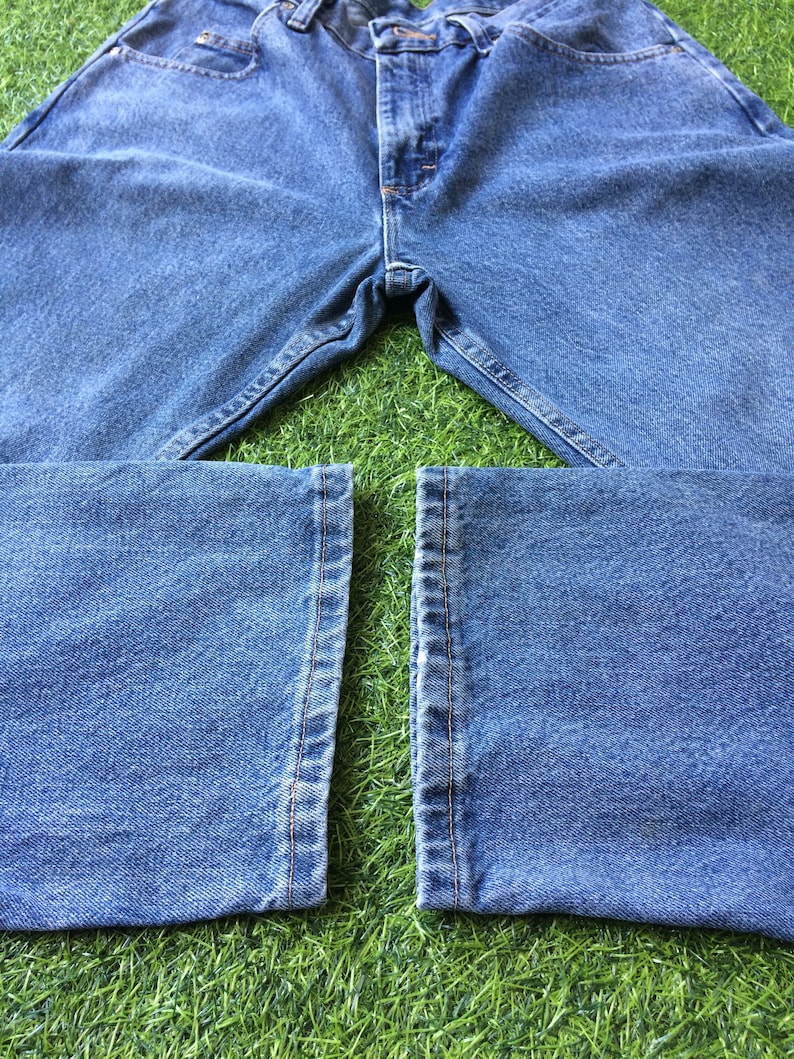 May include: A pair of blue denim jeans laid out on a green grassy surface. The jeans are folded in half, with the legs of the jeans laying flat on the grass.