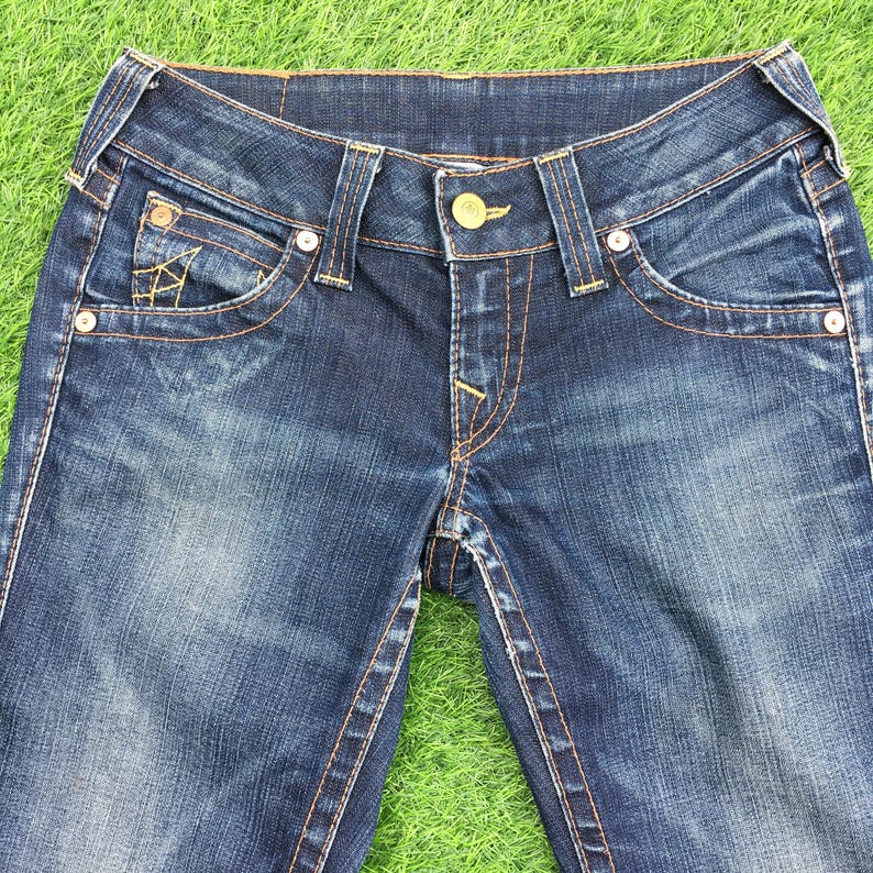 May include: Close-up of blue denim jeans with a button closure and pockets. The jeans have a faded wash and orange stitching. The front pocket features a decorative design. The jeans are displayed on a green surface.