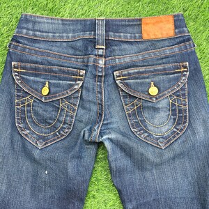 May include: Close-up of the back of blue denim jeans. The jeans have two back pockets with gold stitching and gold button closures. A brown leather patch is at the waistband.