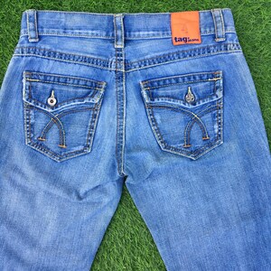 May include: Light blue denim jeans with decorative stitching and unique pocket designs. The back of the jeans features two pockets with curved stitching and metal button details. An orange tag with the text "tag jeans" is visible.