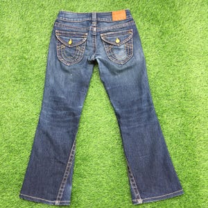 May include: Blue denim jeans with a bootcut leg. The jeans feature two back pockets with decorative stitching and gold-colored button closures. A leather patch is visible at the waistband. The jeans are laid out on a green surface.