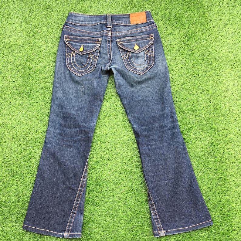 May include: Blue denim jeans with a bootcut leg. The jeans feature two back pockets with decorative stitching and gold-colored button closures. A leather patch is visible at the waistband. The jeans are laid out on a green surface.