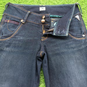 Op de afbeelding: Donkerblauwe denim jeans met een knoop- en ritssluiting. De jeans hebben een klassiek vijf-zakken ontwerp met oranje stiksels en een klein rood accent op de voorzak. De taille heeft twee decoratieve knopen en een riemlus.