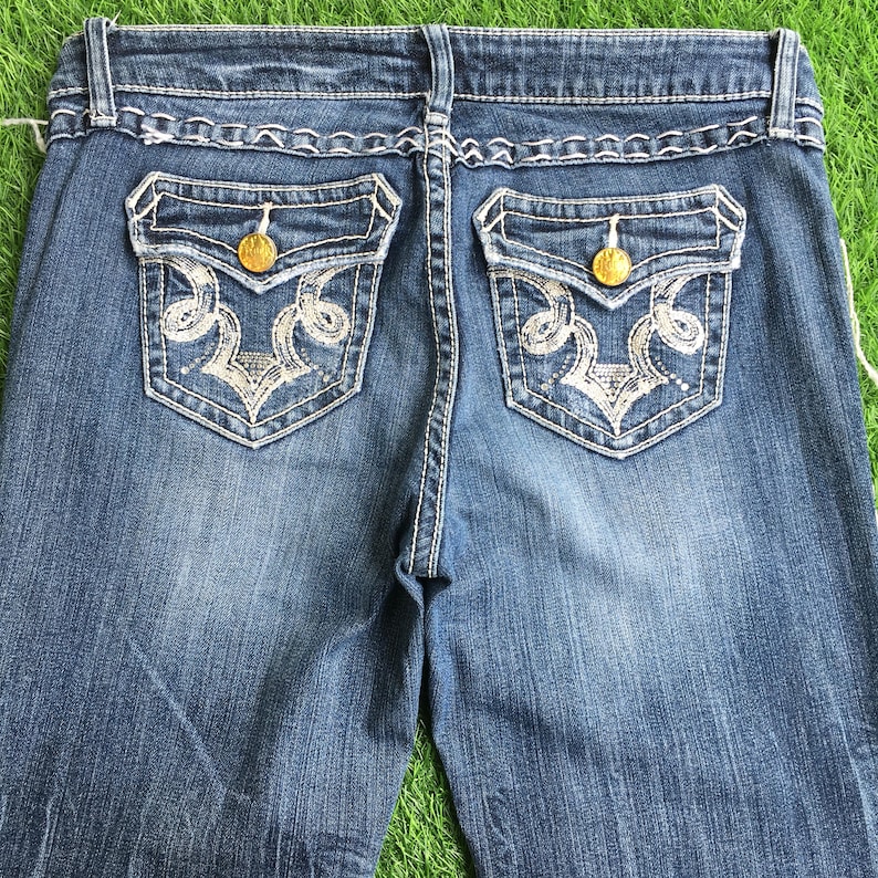 Op de afbeelding: Een paar blauwe denim jeans met geborduurde zakken. De zakken hebben een wit bloemenpatroon en gouden knopen.