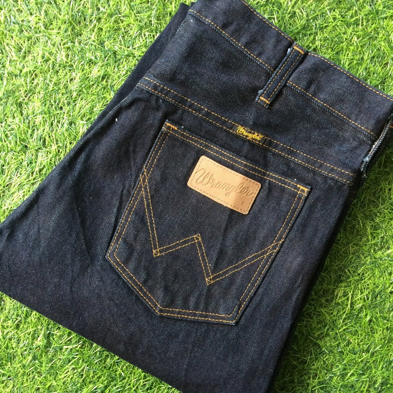Peut inclure: Un jean en denim bleu fonc&eacute; avec une poche arri&egrave;re cousue jaune et une &eacute;tiquette Wrangler en cuir marron.