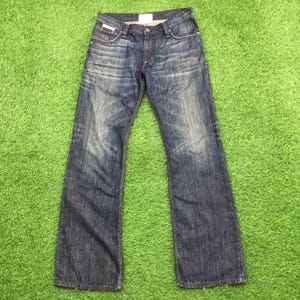 Puede incluir: Un par de vaqueros de mezclilla azul con un lavado descolorido. Los vaqueros tienen un diseño clásico de cinco bolsillos, cierre de botón y cremallera, y una pernera recta. Los vaqueros están planos sobre una superficie verde.