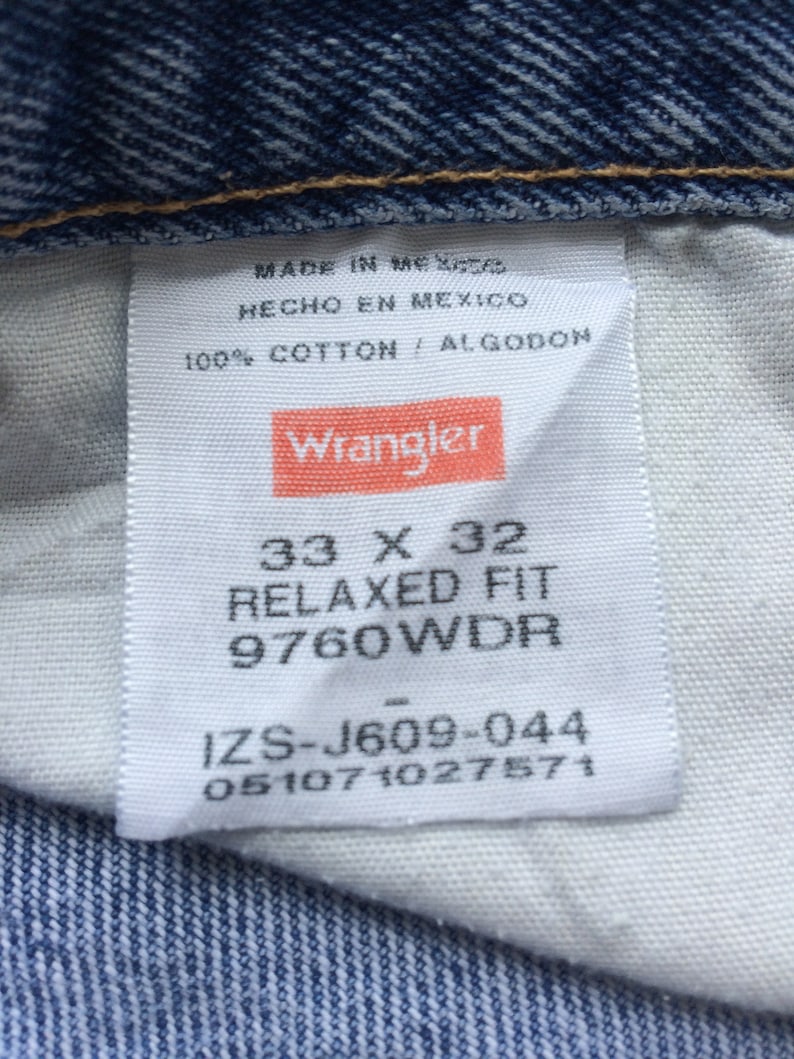 Pode incluir: Uma etiqueta branca com texto preto e um logotipo vermelho Wrangler. A etiqueta diz "Made in Mexico", "Hecho en Mexico", "100% Cotton / Algodon", "33 x 32", "Relaxed Fit", "9760WDR", "IZS-J609-044" e "051071027571".