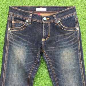 May include: Dark wash denim jeans with a classic five-pocket design. The jeans feature a button closure, rivets, and contrasting gold stitching. The jeans are displayed on a green surface.