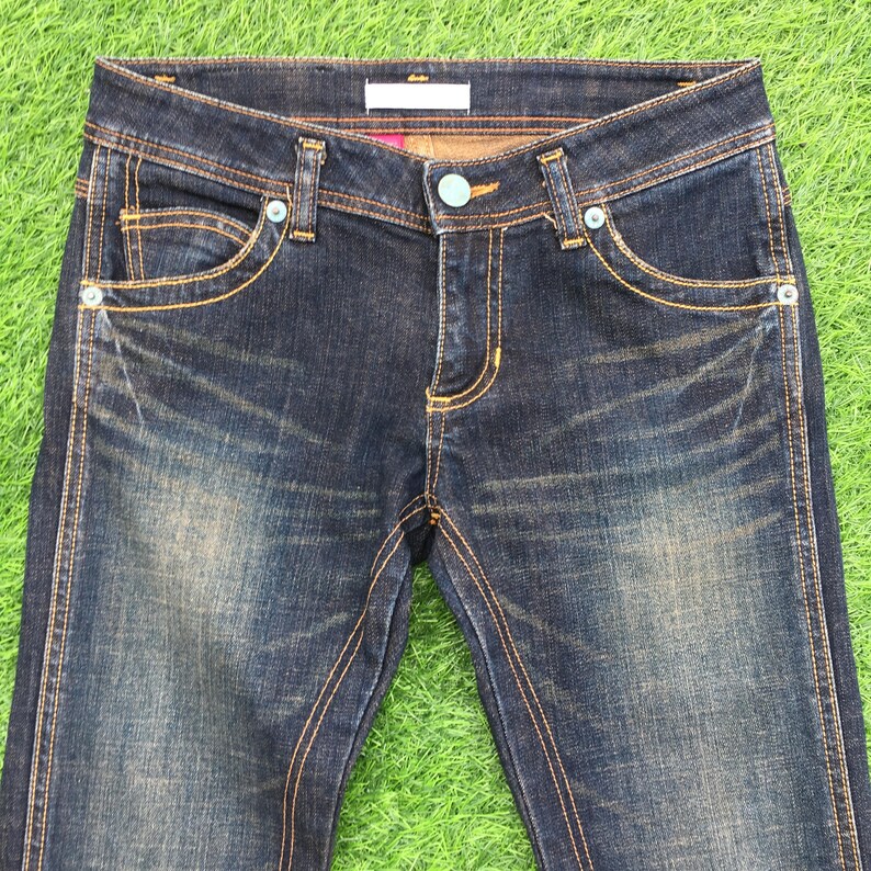 May include: Dark wash denim jeans with a classic five-pocket design. The jeans feature a button closure, rivets, and contrasting gold stitching. The jeans are displayed on a green surface.