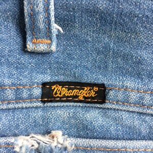 May include: Close-up of a blue denim fabric with a brown leather patch and a black label that reads "Wrangler" in gold lettering.