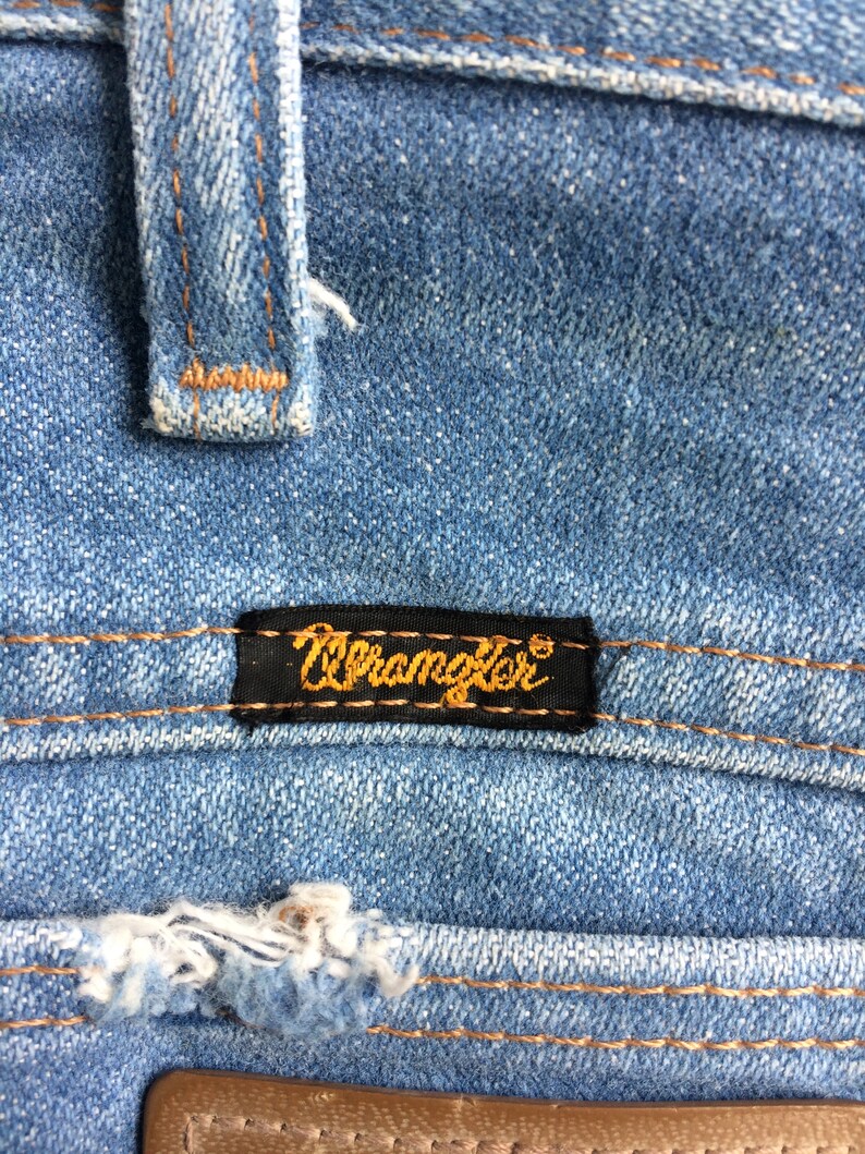 May include: Close-up of a blue denim fabric with a brown leather patch and a black label that reads "Wrangler" in gold lettering.