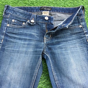 May include: Close-up of blue denim jeans with a partially unzipped fly. The jeans have a classic five-pocket design, with decorative rivets and stitching. The brand name "CECIL McBEE" is visible on the waistband.