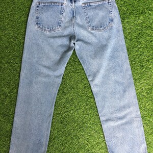 May include: A pair of light blue denim jeans with a brown leather patch on the back waistband that says "Wrangler" in white text. The jeans have a straight leg fit and a frayed hem.