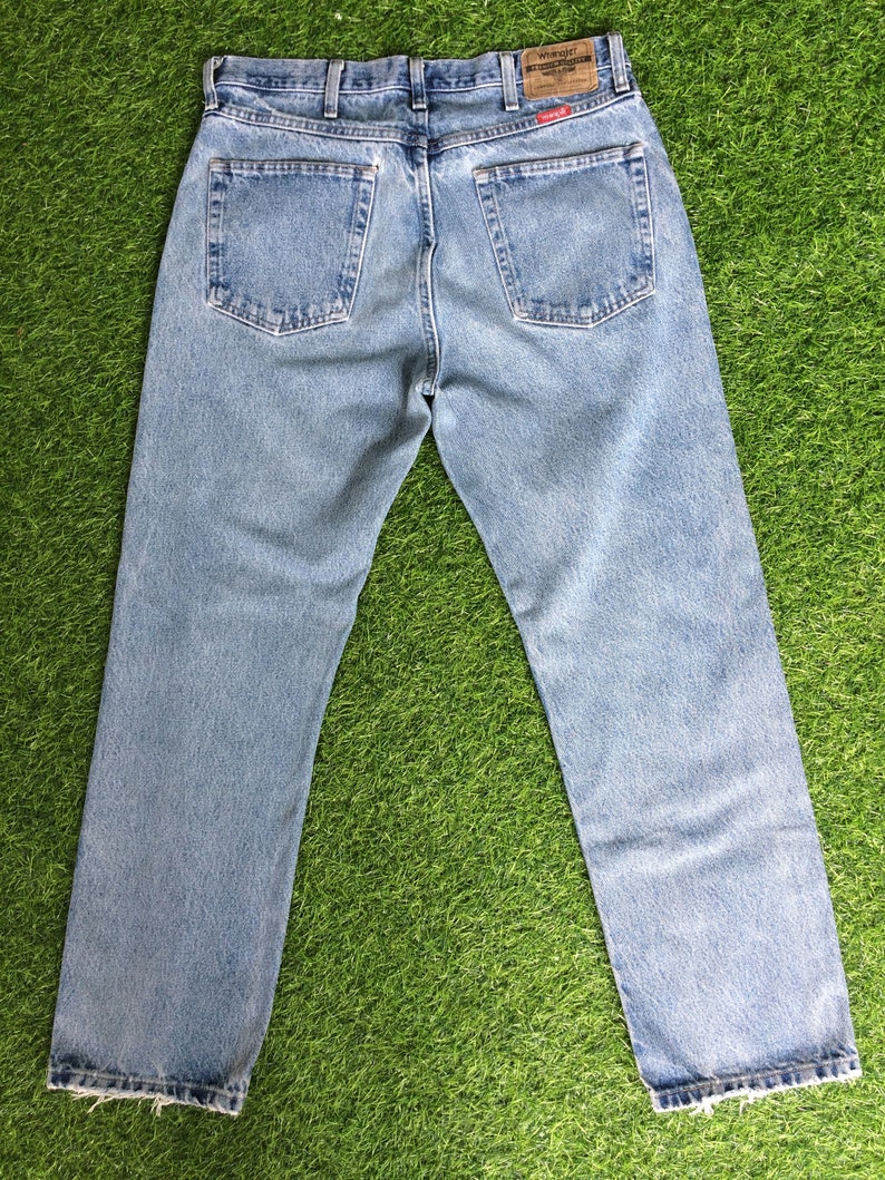 May include: A pair of light blue denim jeans with a brown leather patch on the back waistband that says "Wrangler" in white text. The jeans have a straight leg fit and a frayed hem.
