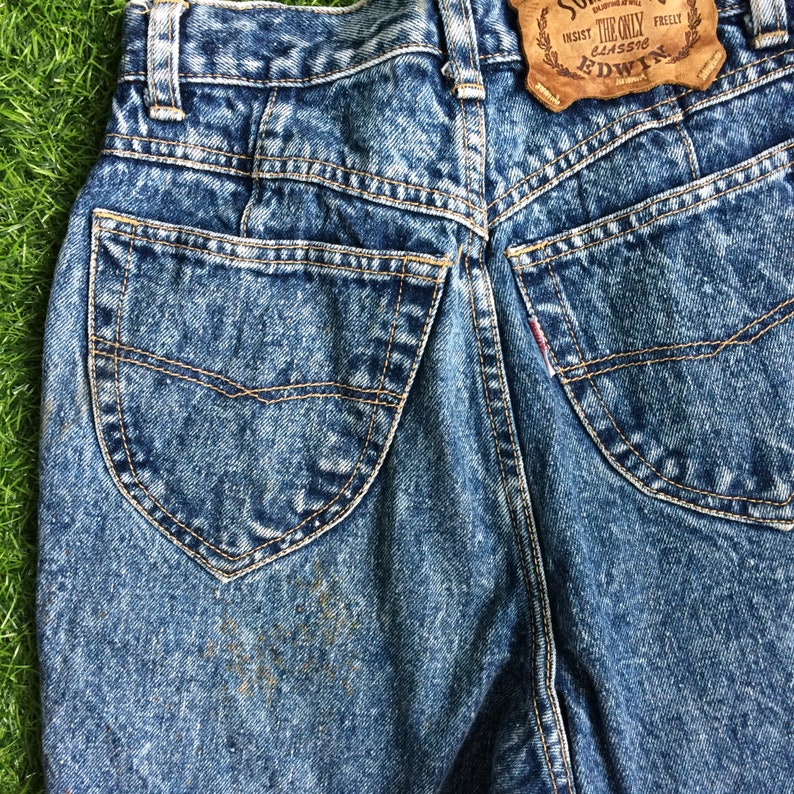 May include: Blue denim jeans with a faded wash and a back pocket with a stitched design. The jeans have a brown leather patch with the text "The Only Classic" and "Edwin" in white.