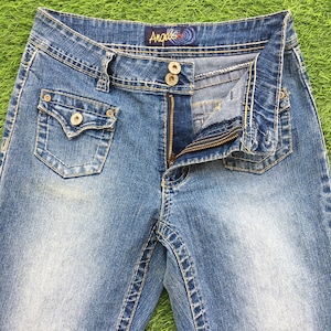 May include: A pair of blue denim jeans with a zipper fly and a button closure. The jeans have a faded look and have a small pocket on the front of each leg. The jeans have the brand name "Angelle" on the waistband.