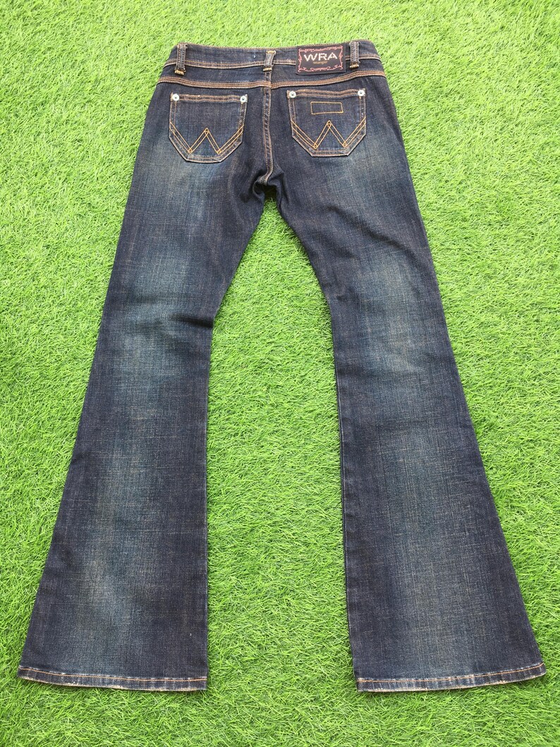 May include: Dark wash denim jeans with a flared leg design. The jeans feature classic five-pocket styling, with decorative stitching on the back pockets and the brand name "WRA" on the waistband.