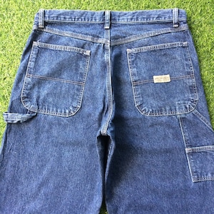 May include: A pair of dark blue denim carpenter jeans with multiple pockets and a small label on the back right pocket that reads "Wrangler Jeans Co."