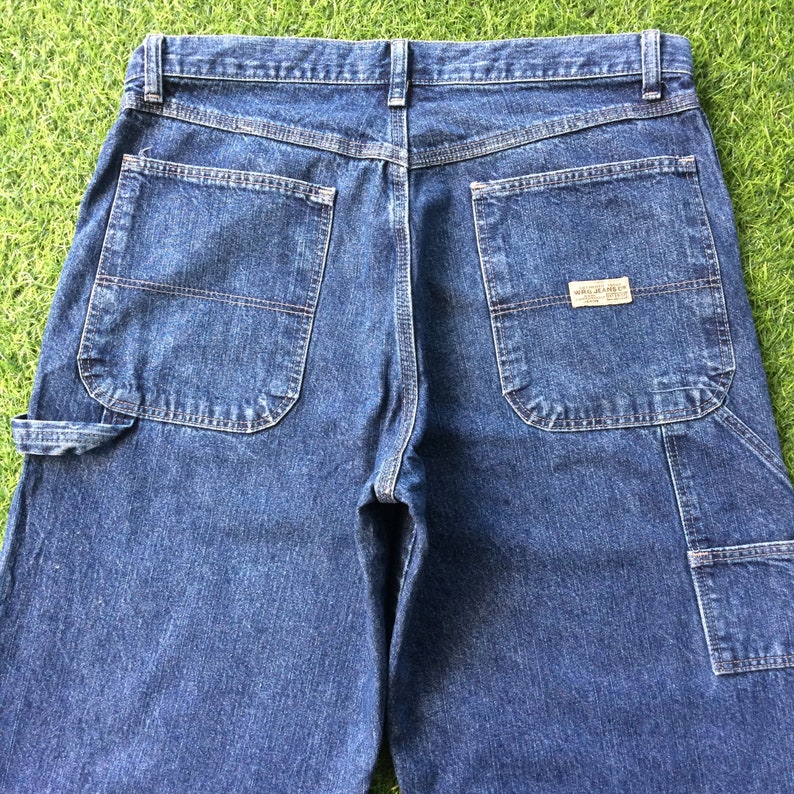 May include: A pair of dark blue denim carpenter jeans with multiple pockets and a small label on the back right pocket that reads "Wrangler Jeans Co."
